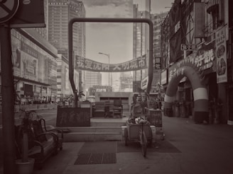 A black and white urban scene featuring tall buildings, a street with various signs in Chinese, and a large archway with promotional banners. Several pieces of furniture and a cart are placed in the foreground, along with potted plants and a variety of small shops on the side.