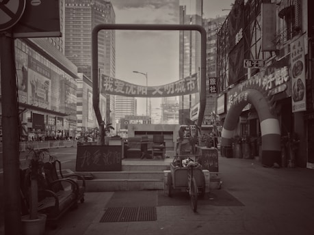 A black and white urban scene featuring tall buildings, a street with various signs in Chinese, and a large archway with promotional banners. Several pieces of furniture and a cart are placed in the foreground, along with potted plants and a variety of small shops on the side.