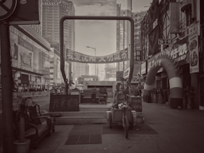 A black and white urban scene featuring tall buildings, a street with various signs in Chinese, and a large archway with promotional banners. Several pieces of furniture and a cart are placed in the foreground, along with potted plants and a variety of small shops on the side.