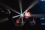 A musician stands on stage under bright spotlight beams, playing a Nord Stage 3 keyboard. Another musician with a guitar is in the background. The stage has musical equipment like a guitar case and water bottles. A few silhouettes of audience members are visible in front.