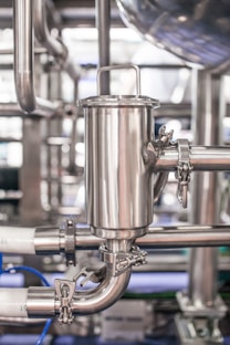 A close-up view of industrial stainless steel piping with clamps and connectors. The machinery appears to be part of a larger industrial system, possibly involving liquid or gas transport. The metal surface has a metallic sheen, and there are several interlocking pipe sections.