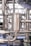 A close-up view of industrial stainless steel piping with clamps and connectors. The machinery appears to be part of a larger industrial system, possibly involving liquid or gas transport. The metal surface has a metallic sheen, and there are several interlocking pipe sections.