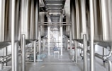 Industrial setting featuring large metallic tanks and pipelines, with a central walkway made of metal grating and smaller machinery visible near the floor.