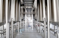 Industrial setting featuring large metallic tanks and pipelines, with a central walkway made of metal grating and smaller machinery visible near the floor.