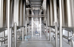 Industrial setting featuring large metallic tanks and pipelines, with a central walkway made of metal grating and smaller machinery visible near the floor.