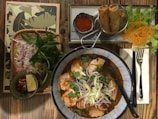 An appealing Vietnamese meal setup on a wooden table includes a bowl of spicy noodle soup garnished with fresh herbs and sliced onions. Accompanying the soup are fried spring rolls served with a small bowl of dipping sauce. Fresh lettuce, carrots, and additional herbs are placed to the side along with a small bowl containing lime and chili.