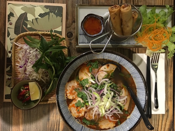 An appealing Vietnamese meal setup on a wooden table includes a bowl of spicy noodle soup garnished with fresh herbs and sliced onions. Accompanying the soup are fried spring rolls served with a small bowl of dipping sauce. Fresh lettuce, carrots, and additional herbs are placed to the side along with a small bowl containing lime and chili.
