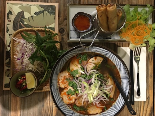 An appealing Vietnamese meal setup on a wooden table includes a bowl of spicy noodle soup garnished with fresh herbs and sliced onions. Accompanying the soup are fried spring rolls served with a small bowl of dipping sauce. Fresh lettuce, carrots, and additional herbs are placed to the side along with a small bowl containing lime and chili.