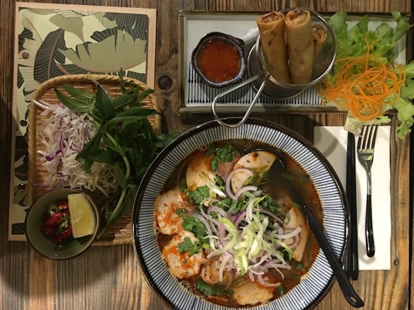 An appealing Vietnamese meal setup on a wooden table includes a bowl of spicy noodle soup garnished with fresh herbs and sliced onions. Accompanying the soup are fried spring rolls served with a small bowl of dipping sauce. Fresh lettuce, carrots, and additional herbs are placed to the side along with a small bowl containing lime and chili.