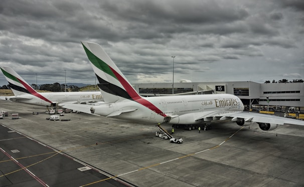 Two large commercial airplanes with Emirates branding are parked at an airport gate under a cloudy sky. The aircraft are connected to jet bridges, and various ground service vehicles and personnel are positioned around them. The scene conveys a typical airport setting with overcast weather.
