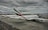 Two large commercial airplanes with Emirates branding are parked at an airport gate under a cloudy sky. The aircraft are connected to jet bridges, and various ground service vehicles and personnel are positioned around them. The scene conveys a typical airport setting with overcast weather.