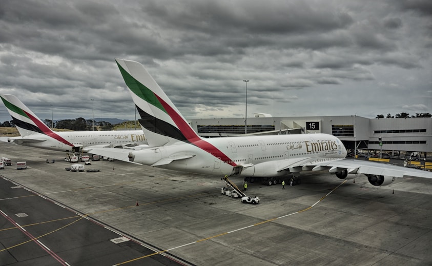 Two large commercial airplanes with Emirates branding are parked at an airport gate under a cloudy sky. The aircraft are connected to jet bridges, and various ground service vehicles and personnel are positioned around them. The scene conveys a typical airport setting with overcast weather.