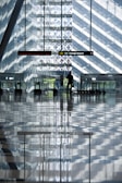 A modern airport terminal featuring large glass walls and strong geometric patterns from the sunlight filtering through. Escalators and information signs are visible, with a lone figure interacting with a digital kiosk. The reflective floor creates an illusion of symmetry and depth.