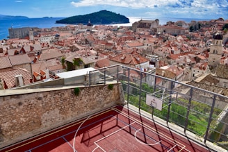 A scenic view of a historic European city with red-tiled roofs and stone buildings, set against a backdrop of the azure sea and distant islands. In the foreground, a rooftop basketball court with a vibrant red surface is enclosed by a metal fence.