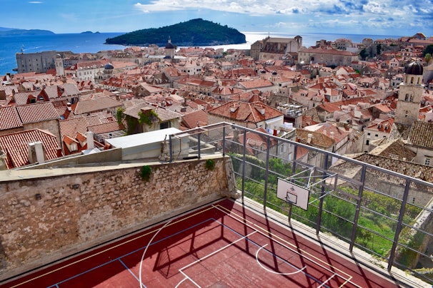 A scenic view of a historic European city with red-tiled roofs and stone buildings, set against a backdrop of the azure sea and distant islands. In the foreground, a rooftop basketball court with a vibrant red surface is enclosed by a metal fence.