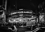 A bustling street food market at night filled with people sitting and enjoying meals under strings of lights. A large sign reading 'Street Food Market' is prominently displayed, with additional text mentioning 'Ben Thanh' and 'Authentic.' Motorbikes are visible in the foreground, indicating a lively and active street scene.