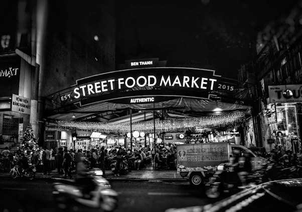 A bustling street food market at night filled with people sitting and enjoying meals under strings of lights. A large sign reading 'Street Food Market' is prominently displayed, with additional text mentioning 'Ben Thanh' and 'Authentic.' Motorbikes are visible in the foreground, indicating a lively and active street scene.
