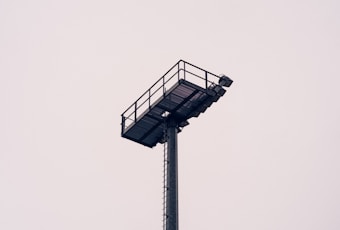 A tall, metallic stadium floodlight tower is set against an overcast sky. The structure is straightforward and industrial, featuring a ladder for maintenance access.