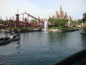 A large theme park scene featuring a fairy tale-style castle with red turrets in the background. In the foreground, there is a large body of water with a fountain spraying up in the air. To the left, there's a red roller coaster winding around its track with a tower structure. The surroundings are lush with greenery and visitors can be seen walking around.