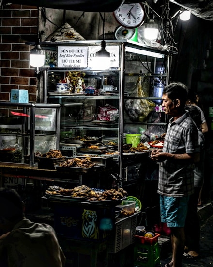 A street food vendor is standing next to a stall filled with trays of cooked food. The stall is lit by overhead lights and contains various items like bowls, pots, and packets. The background features a brick wall and signage advertising noodle soup. A clock hangs on the wall, and there is a cluttered yet inviting atmosphere.