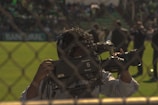 A cameraman operates a professional Panasonic video camera on a sports field, viewed through a chain-link fence. The background features people standing on a grassy area, suggesting a live event or broadcast. The scene is outdoors, with natural lighting.