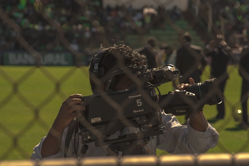 A cameraman operates a professional Panasonic video camera on a sports field, viewed through a chain-link fence. The background features people standing on a grassy area, suggesting a live event or broadcast. The scene is outdoors, with natural lighting.