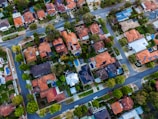 An aerial view of a residential neighborhood with numerous houses lined along curved streets. The houses have varied architectural styles, with many featuring red and blue rooftops, driveways, and surrounding greenery. Cars are parked along the streets and in driveways, and several swimming pools can be seen in some backyards.