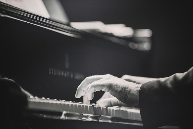 A close-up of a person's hands playing the piano. The image is in black and white, emphasizing the motion and focus on the keys. The piano is a Steinway & Sons model, showing a sense of elegance and quality.