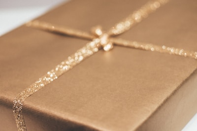A wrapped gift box with a shimmering gold ribbon forming a cross on top. The surface of the box is a matte gold, complementing the ribbon's sparkle.