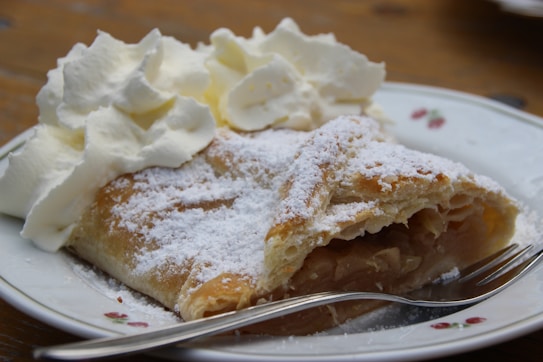 A piece of apple strudel topped with a generous amount of powdered sugar is placed on a decorative plate, accompanied by a swirl of whipped cream. The pastry reveals a layered and flaky texture, with visible apple filling inside, while a fork rests nearby.