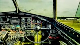 The interior of an airplane cockpit featuring numerous dials, gauges, and control mechanisms. The steering control is prominently centered, and various switches and levers are arranged systematically. Through the windshield, an airfield with parked planes and a hangar is visible against a cloudy sky.