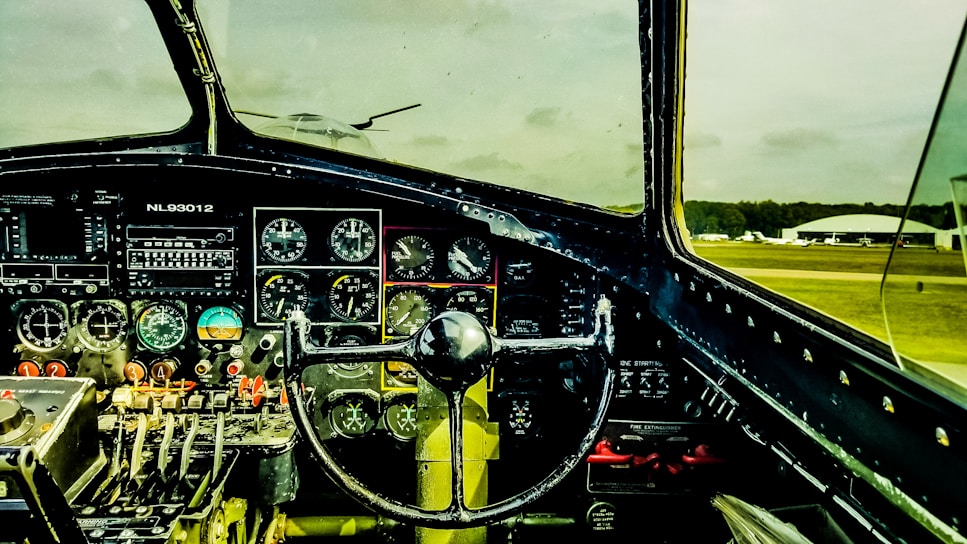 The interior of an airplane cockpit featuring numerous dials, gauges, and control mechanisms. The steering control is prominently centered, and various switches and levers are arranged systematically. Through the windshield, an airfield with parked planes and a hangar is visible against a cloudy sky.