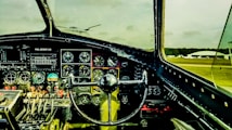 The interior of an airplane cockpit featuring numerous dials, gauges, and control mechanisms. The steering control is prominently centered, and various switches and levers are arranged systematically. Through the windshield, an airfield with parked planes and a hangar is visible against a cloudy sky.