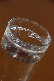 A clear glass filled with water is set against a warm, brown background. Light reflects off the surface of the water, creating gleaming highlights and subtle shadows inside the glass.