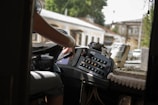 The photo captures the inside of a public transportation vehicle, viewed from the driver's seat. A person is partially visible, focusing on operating the controls and steering wheel. The dashboard is filled with various buttons and a ticket machine. Outside, blurred images of buildings and greenery indicate the vehicle is on a city street.