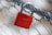 A red padlock adorned with a heart symbol is securely attached to a metal wire fence. The focus is primarily on the lock, with the background being blurred.