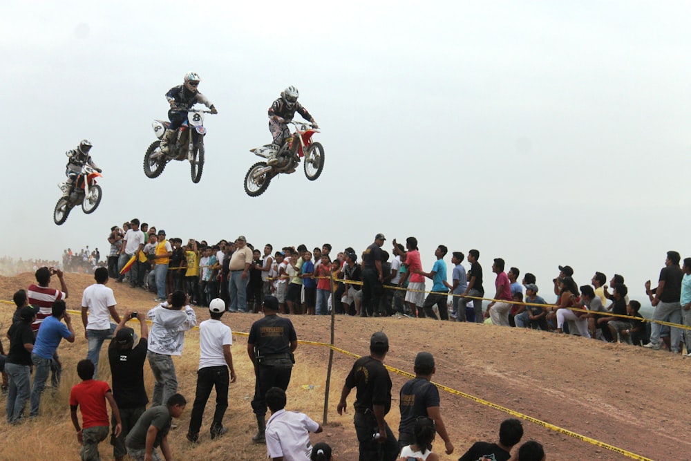 three men riding motocross dirt bikes in mid air near people during day