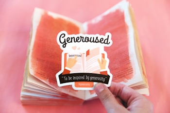 A hand holds a sticker that features an illustration of two hands holding an open book. The sticker includes the word 'Generoused' and the phrase 'To be inspired by generosity' written on it. In the background, a book with pages painted in red is open on a surface with a gradient of red and pink hues.
