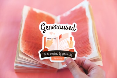 A hand holds a sticker that features an illustration of two hands holding an open book. The sticker includes the word 'Generoused' and the phrase 'To be inspired by generosity' written on it. In the background, a book with pages painted in red is open on a surface with a gradient of red and pink hues.