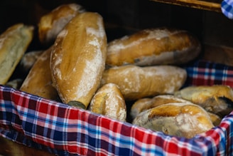 A basket filled with freshly baked loaves of bread. The bread has a golden-brown crust and is arranged on a colorful, checkered cloth.