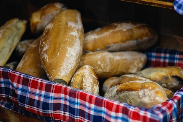 A basket filled with freshly baked loaves of bread. The bread has a golden-brown crust and is arranged on a colorful, checkered cloth.