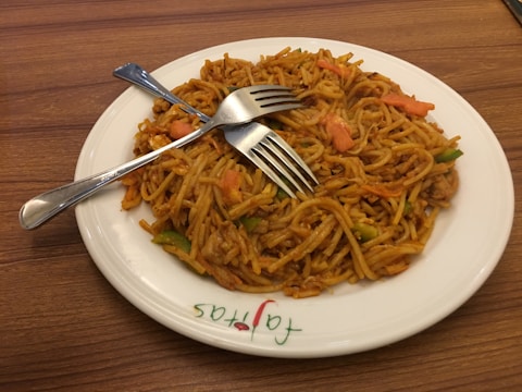 A plate of cooked noodles with vegetables, served on a white plate with two metal forks placed on top. The noodles appear to be mixed with pieces of carrot, green bell pepper, and possibly chicken. The plate rests on a wooden surface.