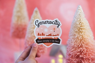 A hand holds a decorative sticker with the word 'Generocity', featuring illustrations of trees and a winter village. In the background, there are cone-shaped, snow-covered artificial trees with a pink and white gradient.