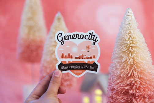 A hand holds a decorative sticker with the word 'Generocity', featuring illustrations of trees and a winter village. In the background, there are cone-shaped, snow-covered artificial trees with a pink and white gradient.