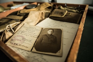 A collection of vintage items including an old black and white photograph of a man in military uniform, an identity card with an official stamp, and various old papers with handwritten text. These items are placed in a wooden box along with a few other military accessories such as straps and pouches.