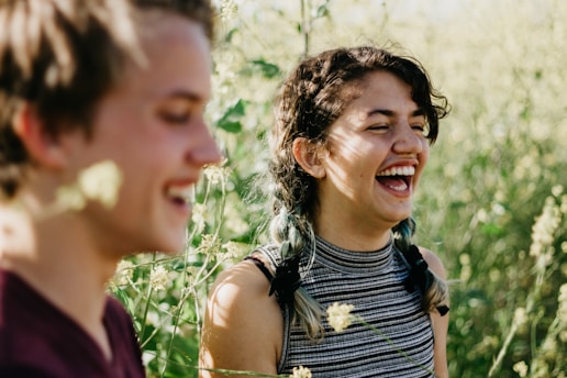 A young woman with dark hair in braids is laughing joyfully in an outdoor setting filled with lush green plants and sunlight. Another person, positioned slightly out of focus and to the side, is also smiling.