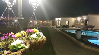 A nighttime scene featuring an outdoor area with decorative lighting, hanging flower baskets, and a pool. The setting includes a well-lit path leading to a building, illuminated windows, and patio furniture.