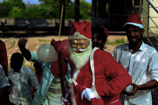 A person dressed as Santa Claus is surrounded by others in festive attire. Santa is holding balloons and carrying a staff, with a background that suggests an outdoor setting.