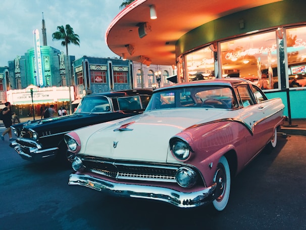 A group of classic muscle cars lined up at a retro drive-in diner under neon lights.