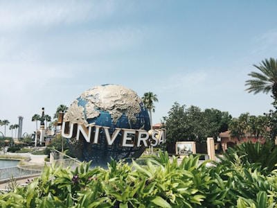 A large globe sculpture with the word 'Universal' in large, metallic letters curves around the front. The structure is surrounded by lush greenery and palm trees under a clear blue sky.